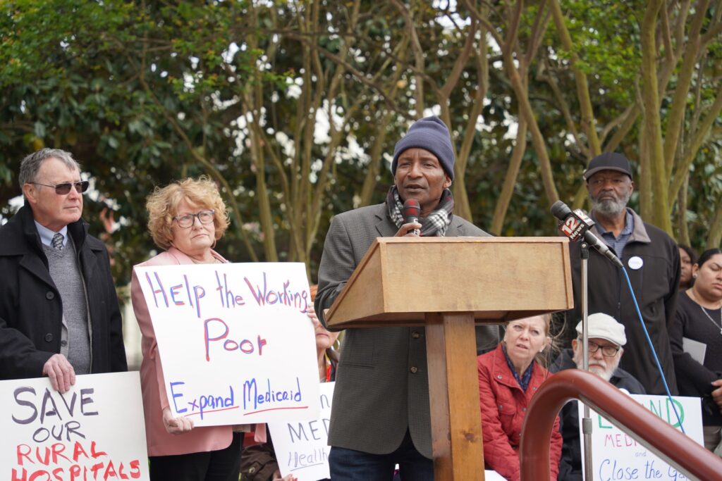 Kenneth King speaks from behind a lectern outside the Alabama State House in Montgomery. He is a Black man wearing a gray suit, a gray toboggan cap and a scarf that is white and black. Standing behind him are numerous supporters of Medicaid expansion, including a white man wearing a black jacket and glasses who holds a sign reading "Save Our Rural Hospitals," and a white woman with red hair and glasses who holds a sign reading "Help the Working Poor: Expand Medicaid."