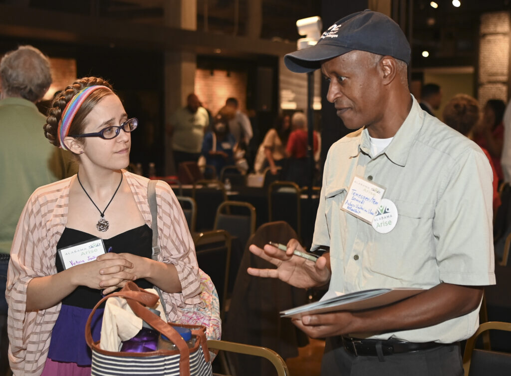 Two Alabama Arise members speak at our 2023 Annual Meeting. On the left is a white woman wearing glasses with a black blouse and a striped pink shirt over it. She has a purse over her shoulder and a bag in front of her. On the right is a Black man wearing a black hat and a cream-colored shirt with an Alabama Arise button. Both are wearing nametags.