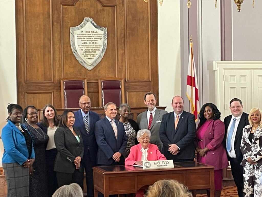 Smiling Alabama Arise staff members and current and former legislators stand behind and to either side of Gov. Kay Ivey, who is seated at a desk in the Old House Chamber in the State Capitol in Montgomery.