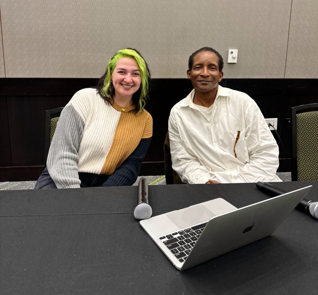 Two Alabama Arise storytellers, Eryn Mullins and Kenneth Tyrone King, smile for a photo while sitting behind a table with a black tablecloth with a laptop and microphone atop it. Eryn is a white woman with dark hair with light green highlights. She is wearing a white and brown sweater with gray and black sleeves. Kenneth is a Black man wearing a white long-sleeved shirt.