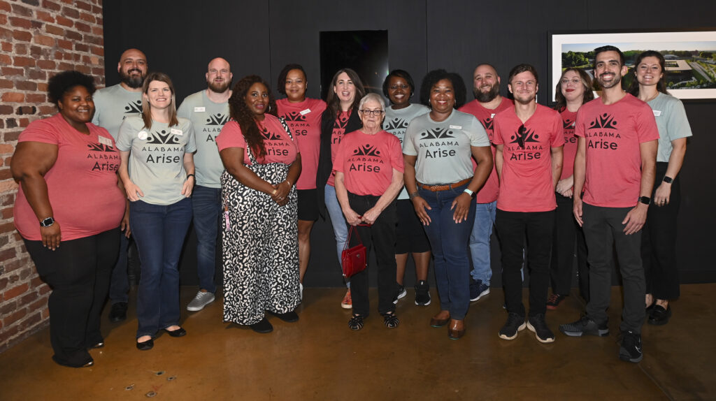 Fifteen Alabama Arise staff members, all wearing either red or green shirts with the Arise logo, stand and smile for a group photo. To their left is a red brick wall, and behind them is a black wall with two framed photographs.