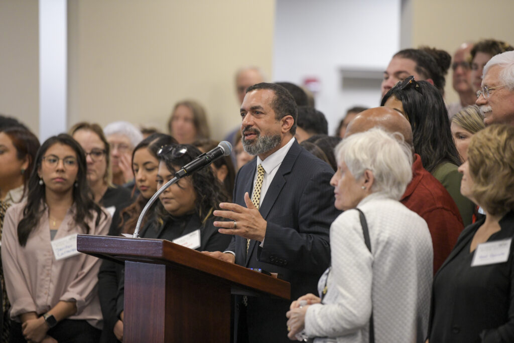 A bearded Black man wearing a black suit jacket, white dress shirt and yellow tie speaks behind a lectern. Alabama Arise supporters stand behind and to either side of him.