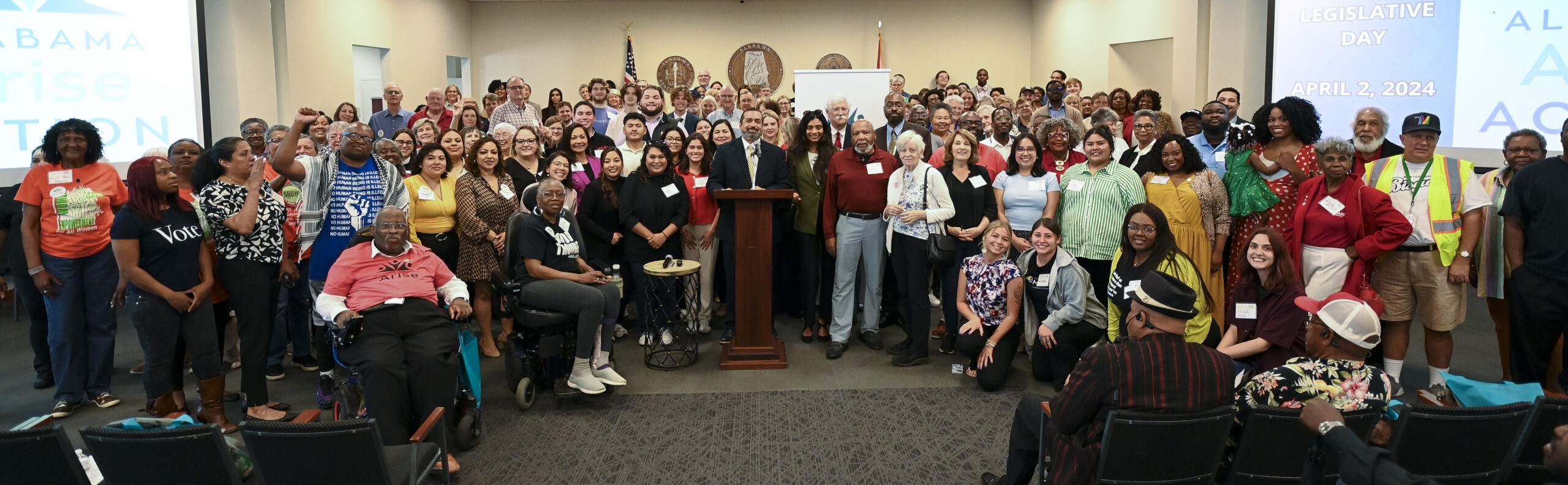 A crowd of hundreds of smiling people gathered behind and to both sides of a lectern inside a committee room in the Alabama State House.