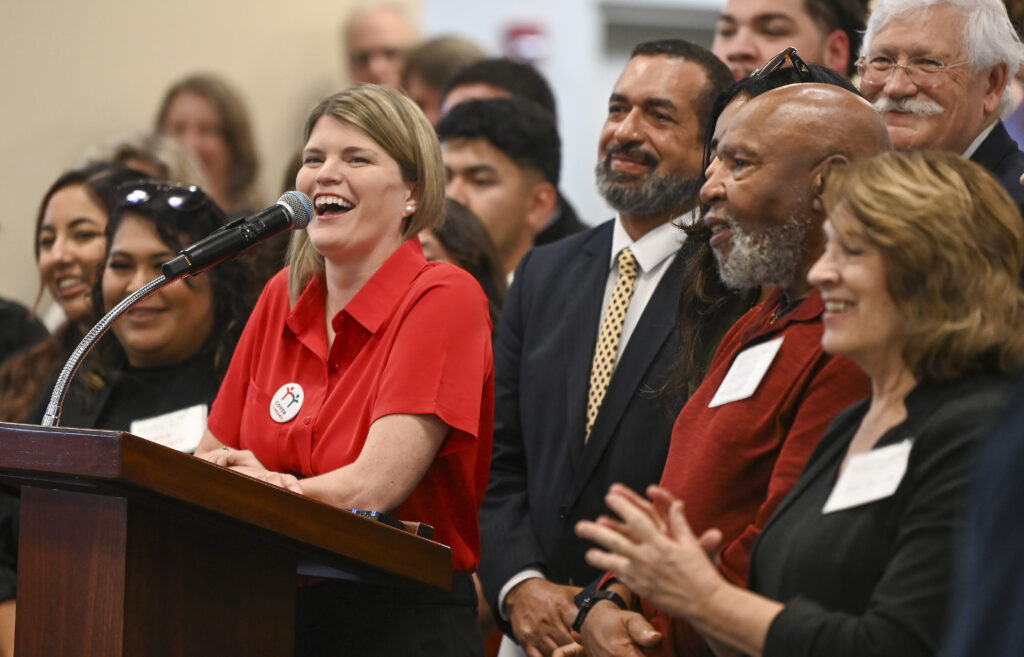 A smiling white woman with blonde hair wearing a light red blouse speaks behind a lectern. Alabama Arise supporters stand behind and to either side of her.