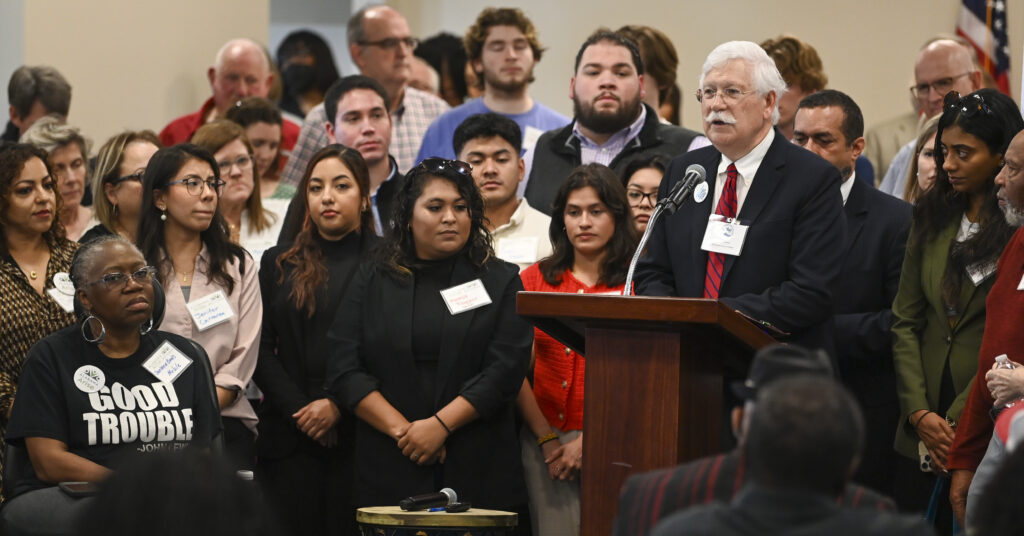 A white man with glasses, white hair and a white mustache stands behind a lectern. He is wearing a black suit jacket, a white dress shirt and a red tie. Alabama Arise supporters stand behind and to either side of him.