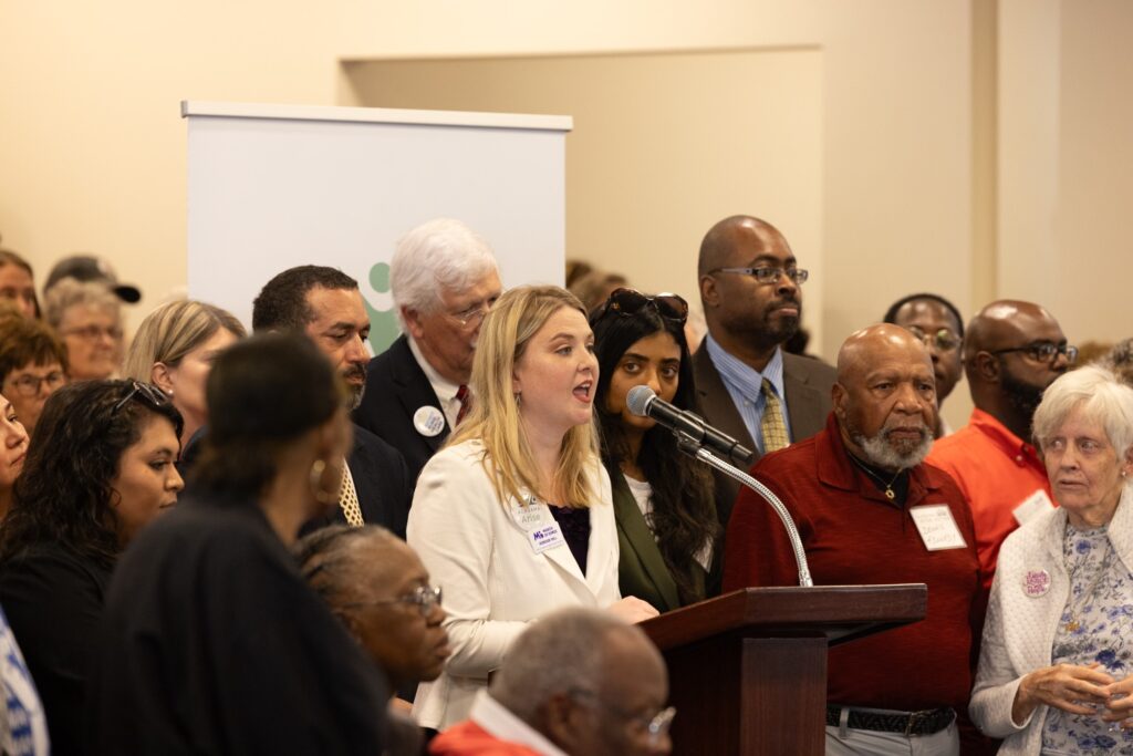 A white woman with blonde hair wearing a white suit jacket and a black blouse speaks behind a lectern. Alabama Arise supporters stand behind and to either side of her.